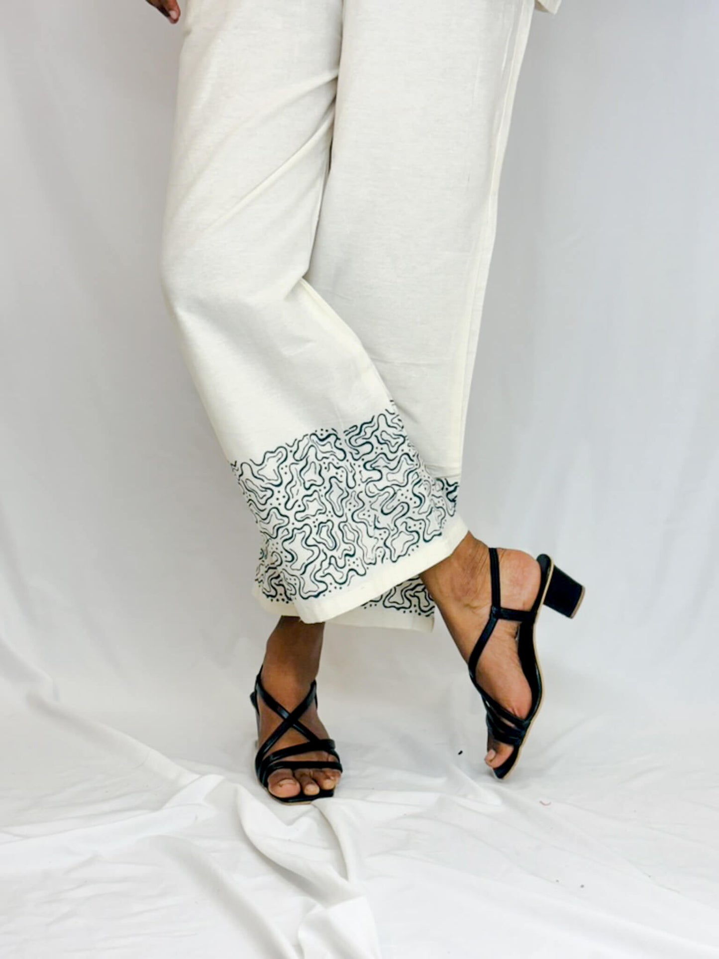 White pants with black patterned hem and black strappy sandals on a white background
