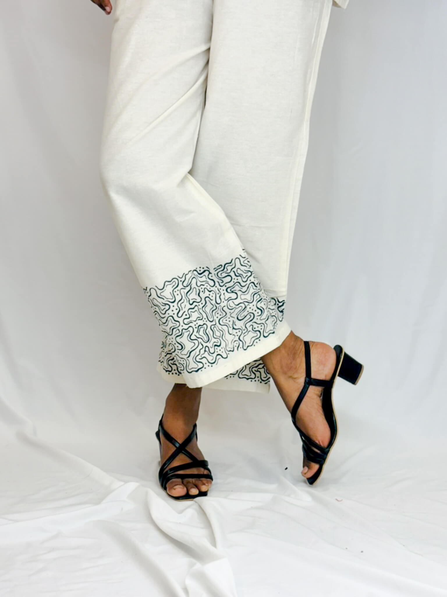 White pants with black patterned hem and black strappy sandals on a white background