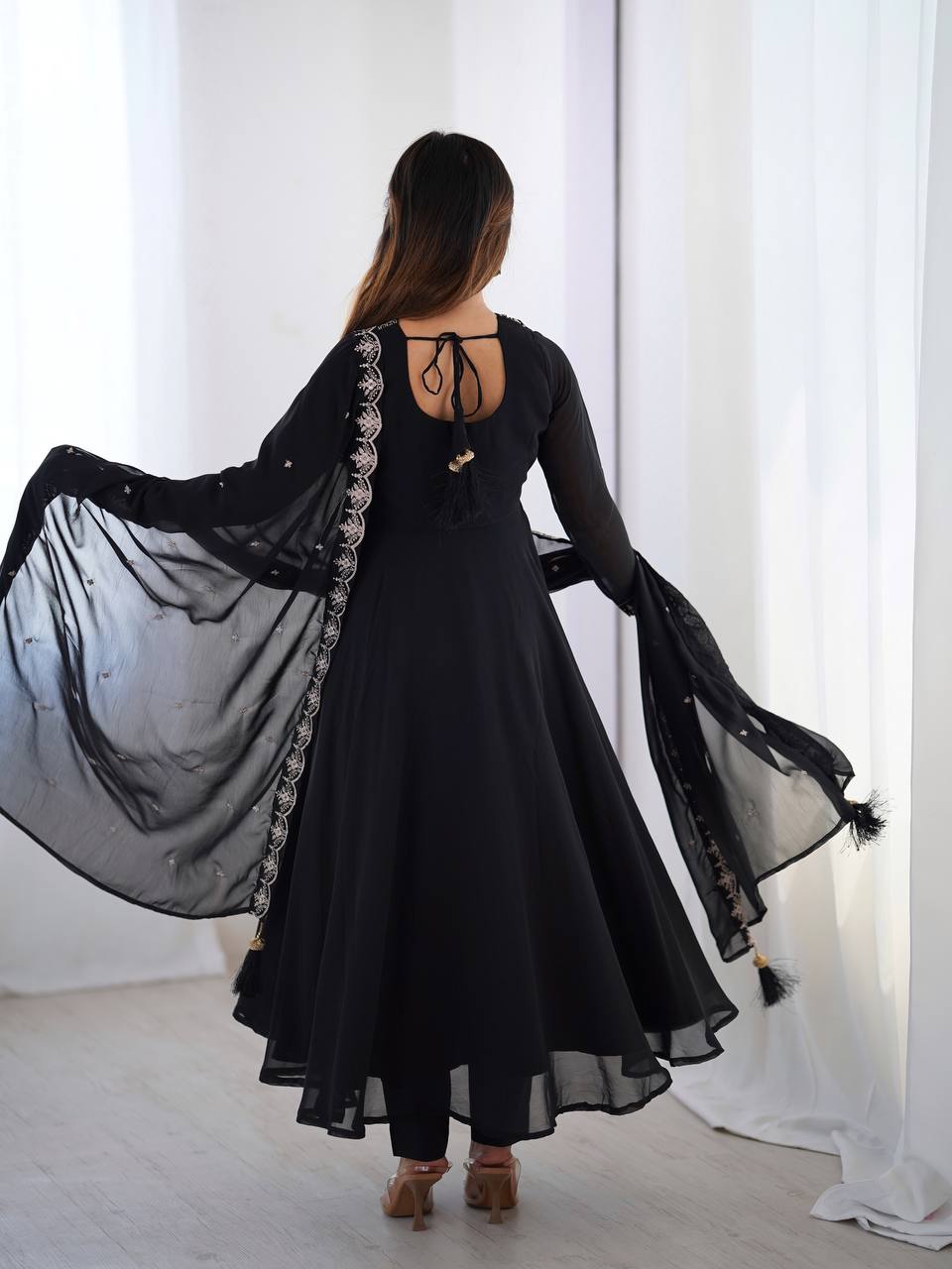 Woman wearing a black dress with sheer sleeves and a long train, standing in front of white curtains.