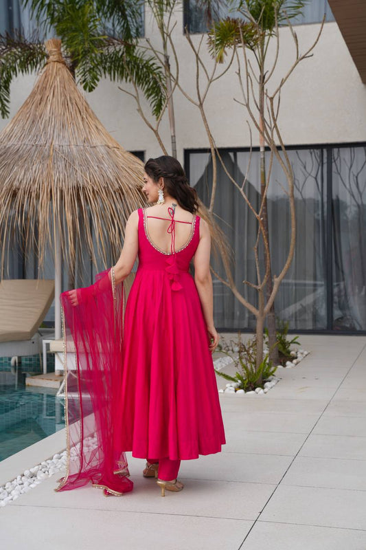 Woman in a pink dress standing by a poolside with a thatched roof structure and trees in the background.