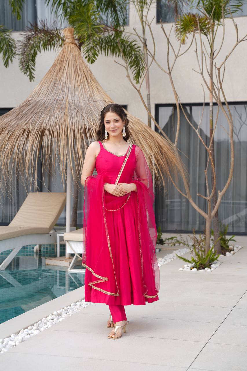 Woman in a pink traditional outfit standing by a poolside with a thatched umbrella and pool chair in the background.