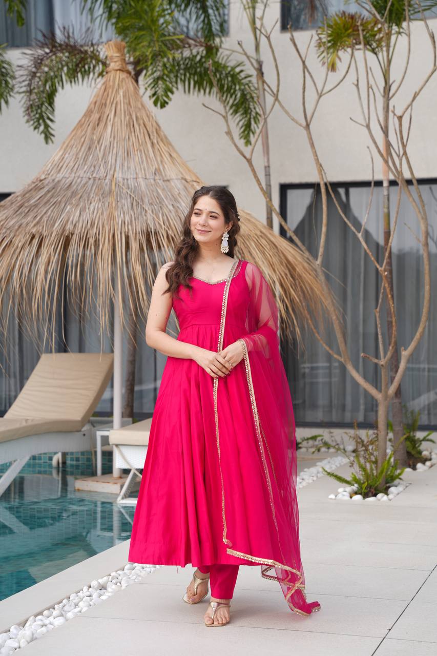 Woman in a pink traditional outfit standing by a poolside with a thatched umbrella and pool in the background.
