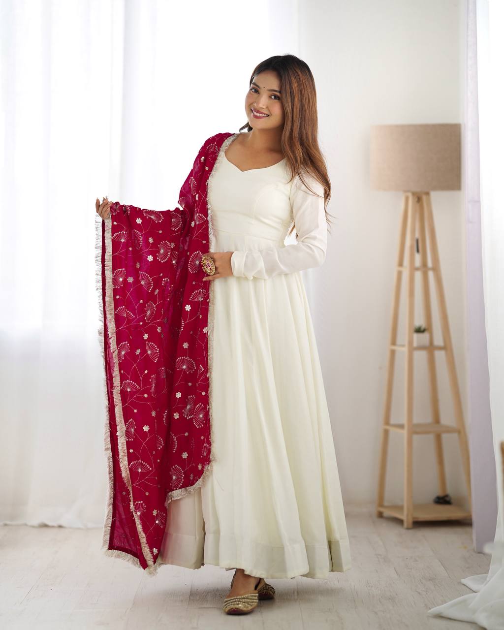 Woman in a white traditional outfit with a red dupatta, standing indoors.