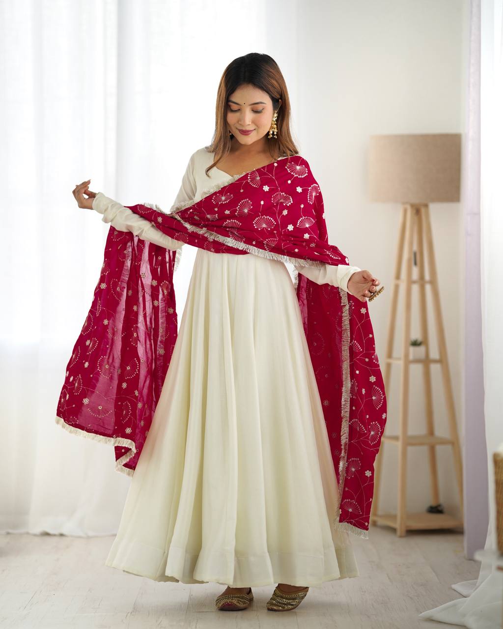 Woman holding a red dupatta with white patterns over a white dress in a bright room.